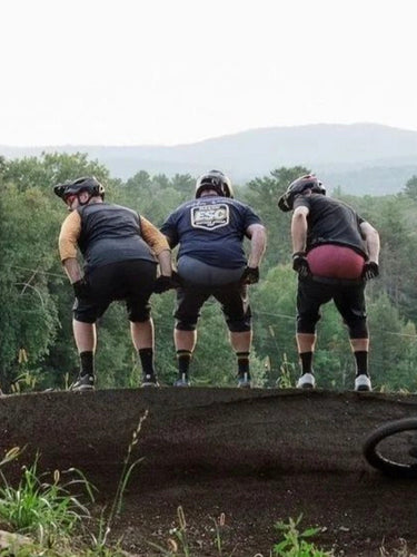 Three mountain bikers preparing to descend a dirt trail with trees and mountains in the background.