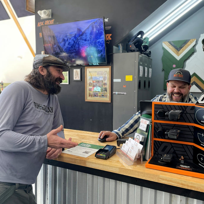 Two men interacting at a counter with a TV and display case in the background.